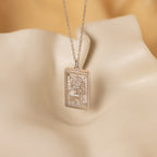The Sun & Moon Tarot Necklace is a gold rectangular pendant featuring engraved sun, moon, diamonds, and animal designs—ideal as statement jewelry—displayed on a cream background.