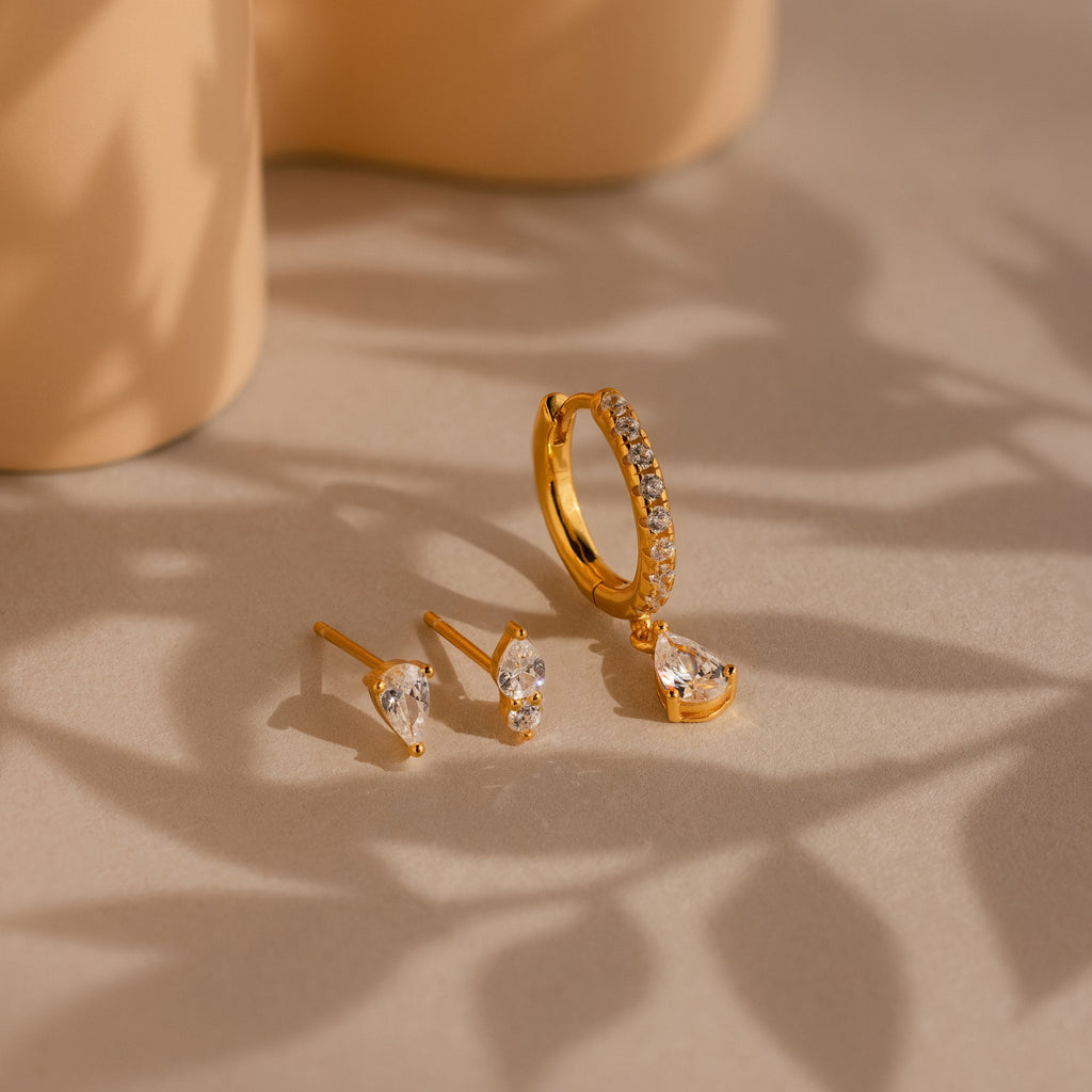 The Diamond Drop Earrings Set, featuring gold pavé hoops with diamonds and matching teardrop stud earrings, is displayed on a beige surface with soft shadows.