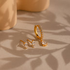 The Diamond Drop Earrings Set, featuring gold pavé hoops with diamonds and matching teardrop stud earrings, is displayed on a beige surface with soft shadows.