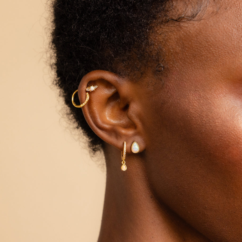 Close-up of a person's ear with three gold cartilage earrings, including a hoop and Opal and Diamond Studs, against a neutral background.
