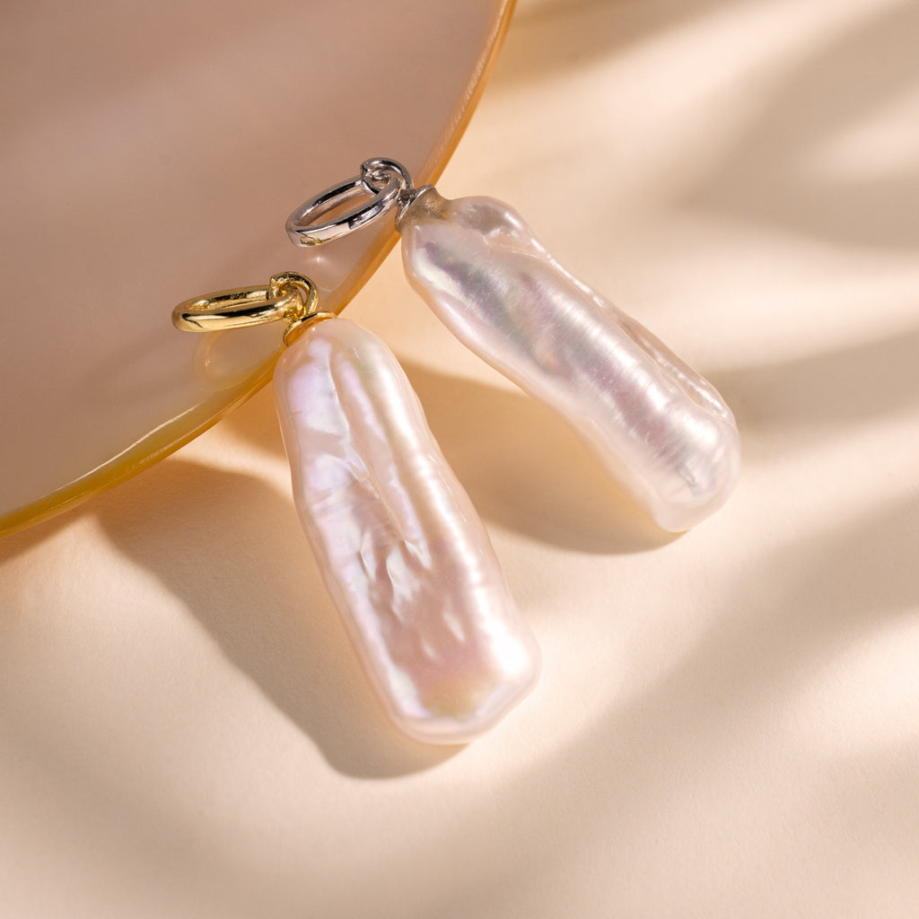 Two Long Baroque Pearl Charms with gold and silver loops rest on a pale, reflective surface, highlighting the unique allure and timeless appeal of these distinctive pearl jewelry pieces.