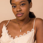 Woman wearing a cream lace camisole and an 18K gold necklace featuring the Long Baroque Pearl Charm, looking at the camera against a beige background.