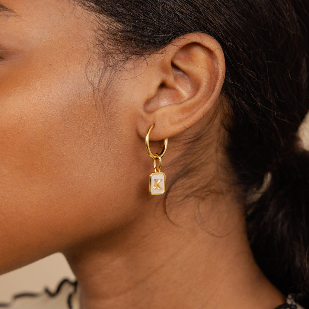 Model wearing a hoop earring with a dangling gold mother of pearl charm n the letter K 