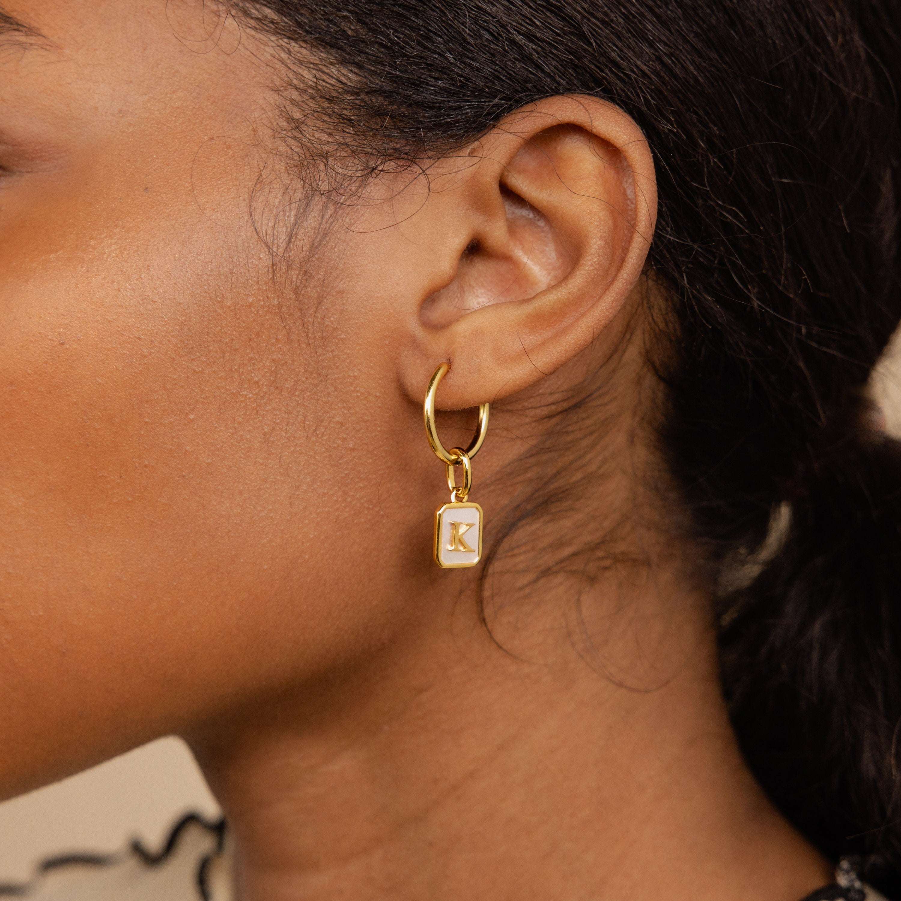 Model wearing a hoop earring with a dangling gold mother of pearl charm n the letter K 