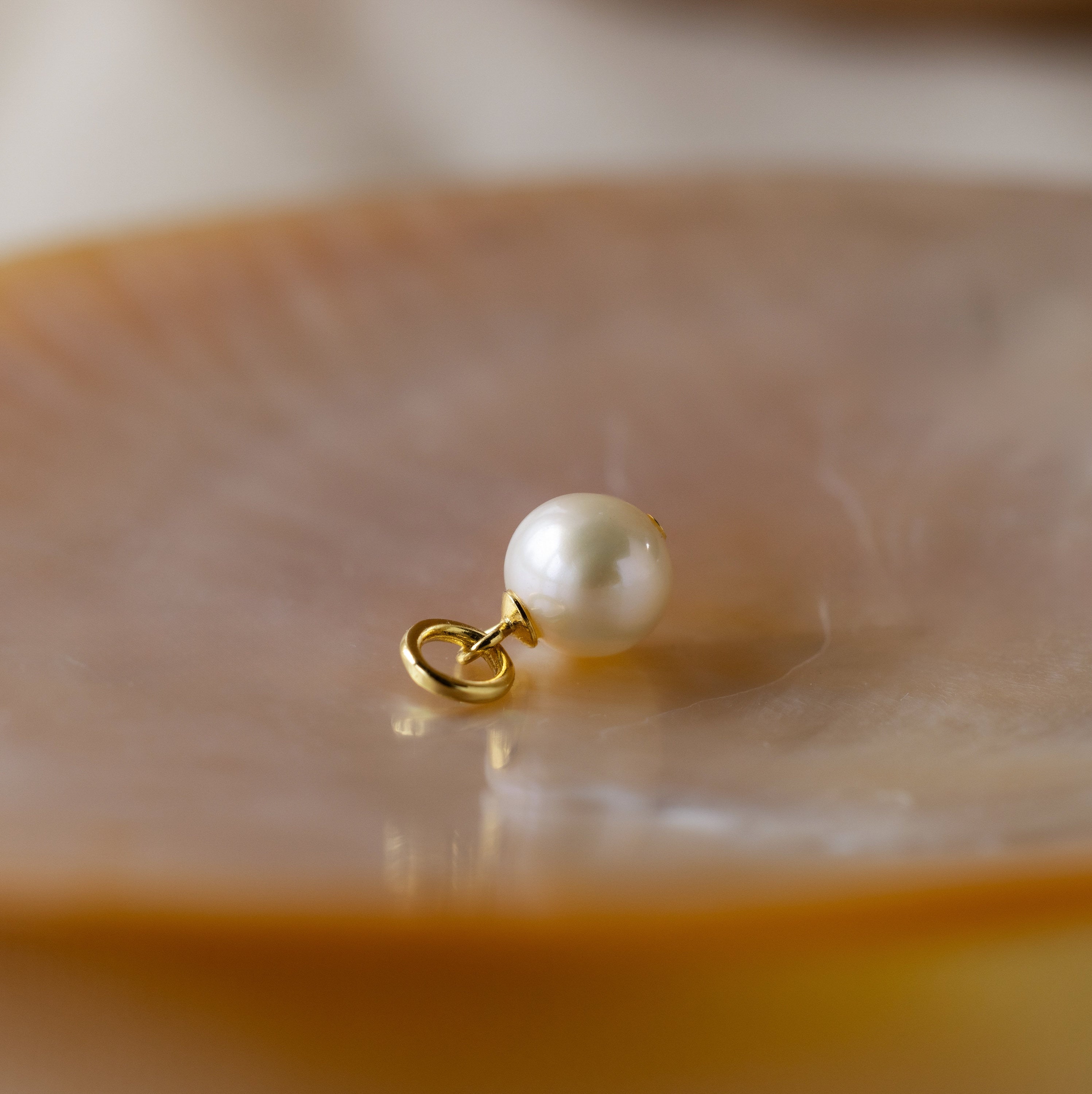 Close up of a minimalist single round white freshwater pearl charm with a gold plated jump ring, resting on a glossy peach toned shell dish. 