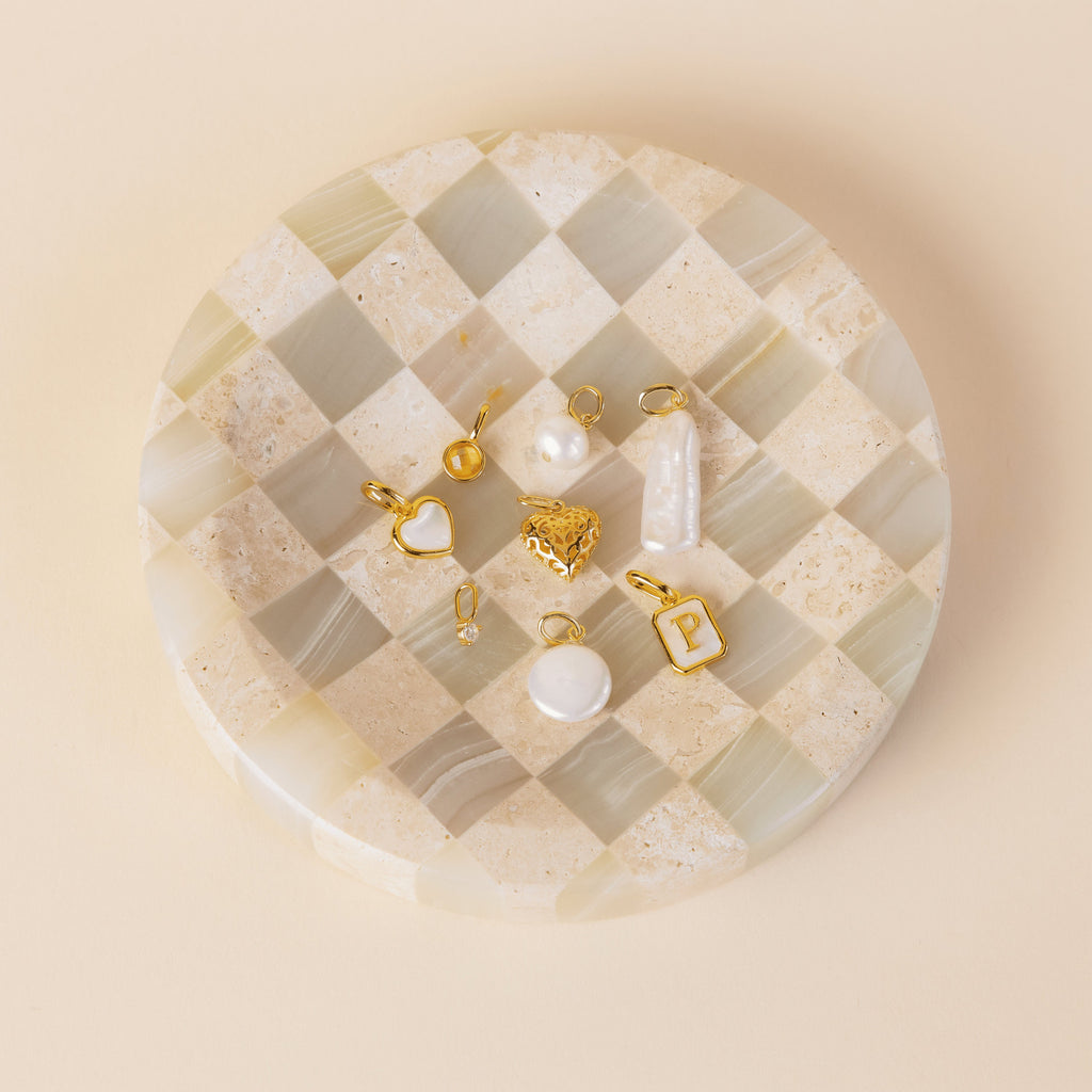 The Classic Pearl Charm in 18K Gold is displayed on a checkered round stone tray against a neutral background.