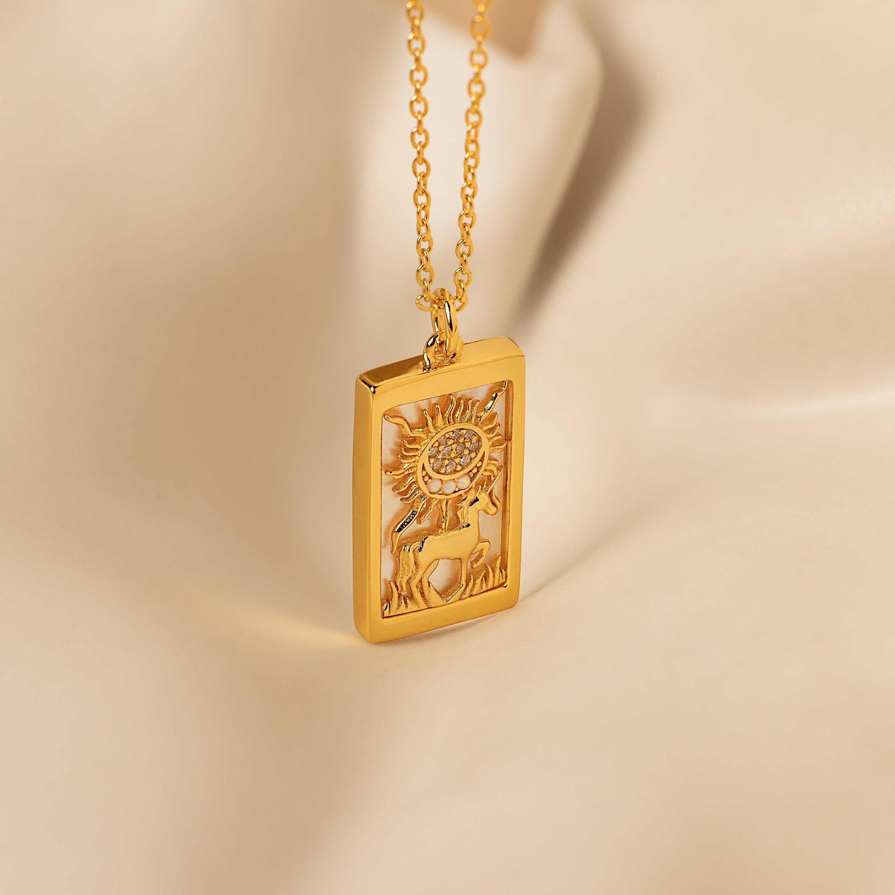 The Sun & Moon Tarot Necklace features a gold rectangular pendant engraved with a sun, moon, rays, and a figure, set against a soft beige background—ideal for those who love statement jewelry.