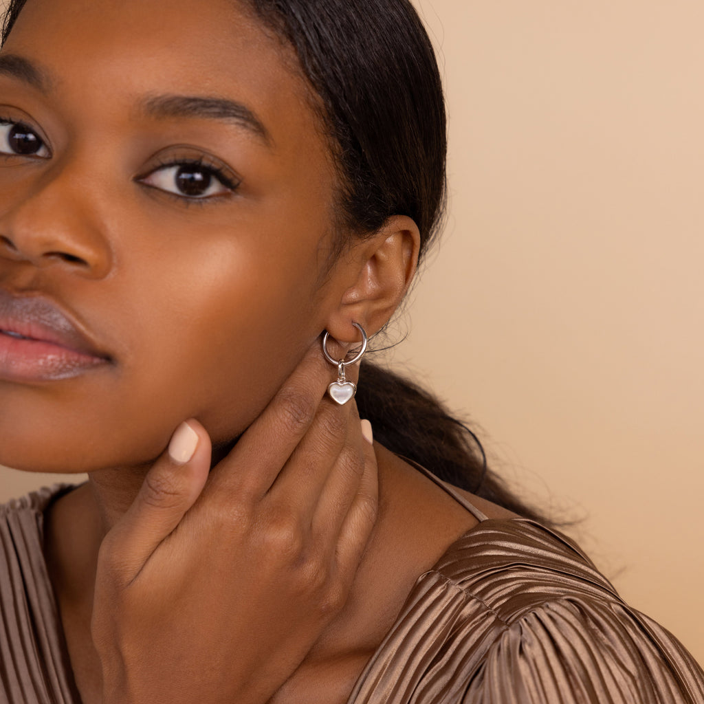 Woman with smooth skin touching her neck wears the Mother of Pearl Heart Charm in Sterling Silver and a pleated brown top, showcasing timeless elegance with vintage-inspired jewelry.