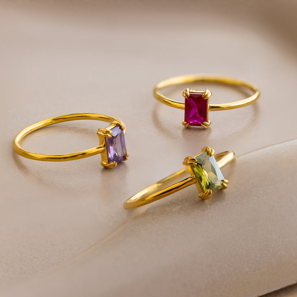 Three Emerald Cut Birthstone Rings, each with red, purple, and green gemstones, are displayed on a soft beige surface—an ideal Christmas gift for someone special.