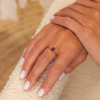 A hand with white painted nails wears the Emerald Cut Birthstone Ring, featuring a small purple gemstone—an elegant and stylish Christmas gift choice.