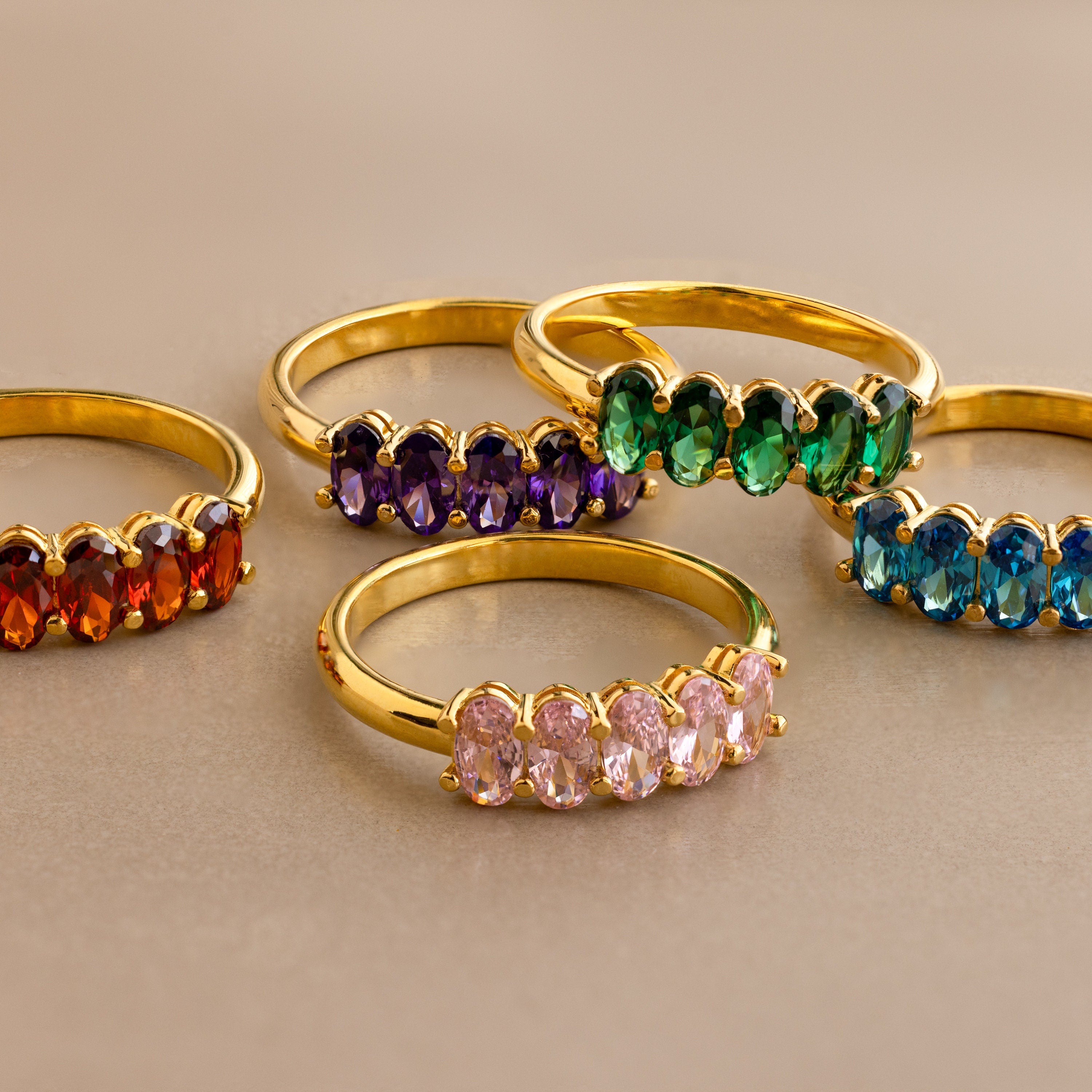 Five Oval Birthstone Rings with red, purple, green, blue, and pink stones are arranged on a beige surface. Each vibrant ring brings colorful style to your jewelry collection.