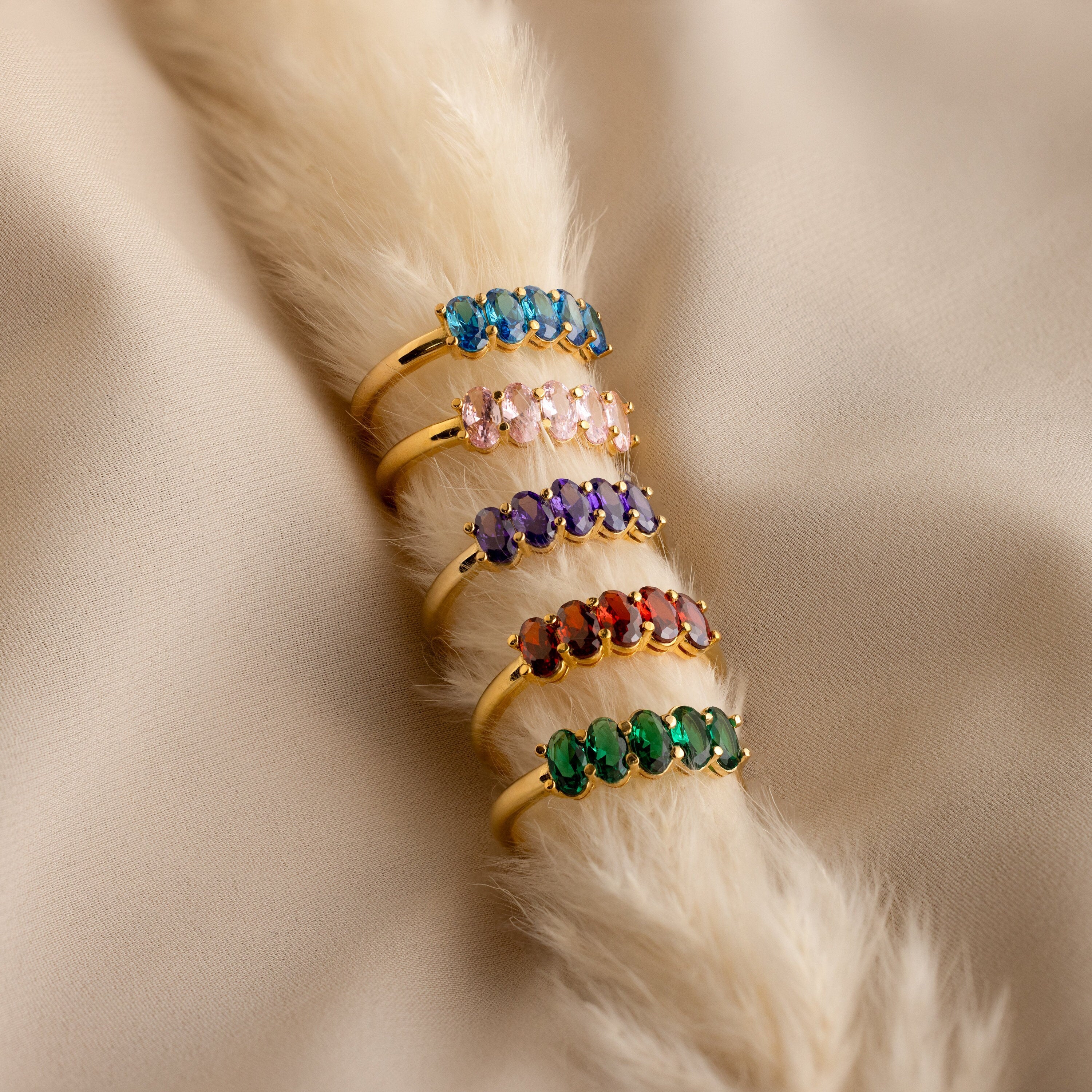 The Oval Birthstone Ring, featuring a personalized gemstone, is displayed among five gold rings on soft beige fabric and faux fur.