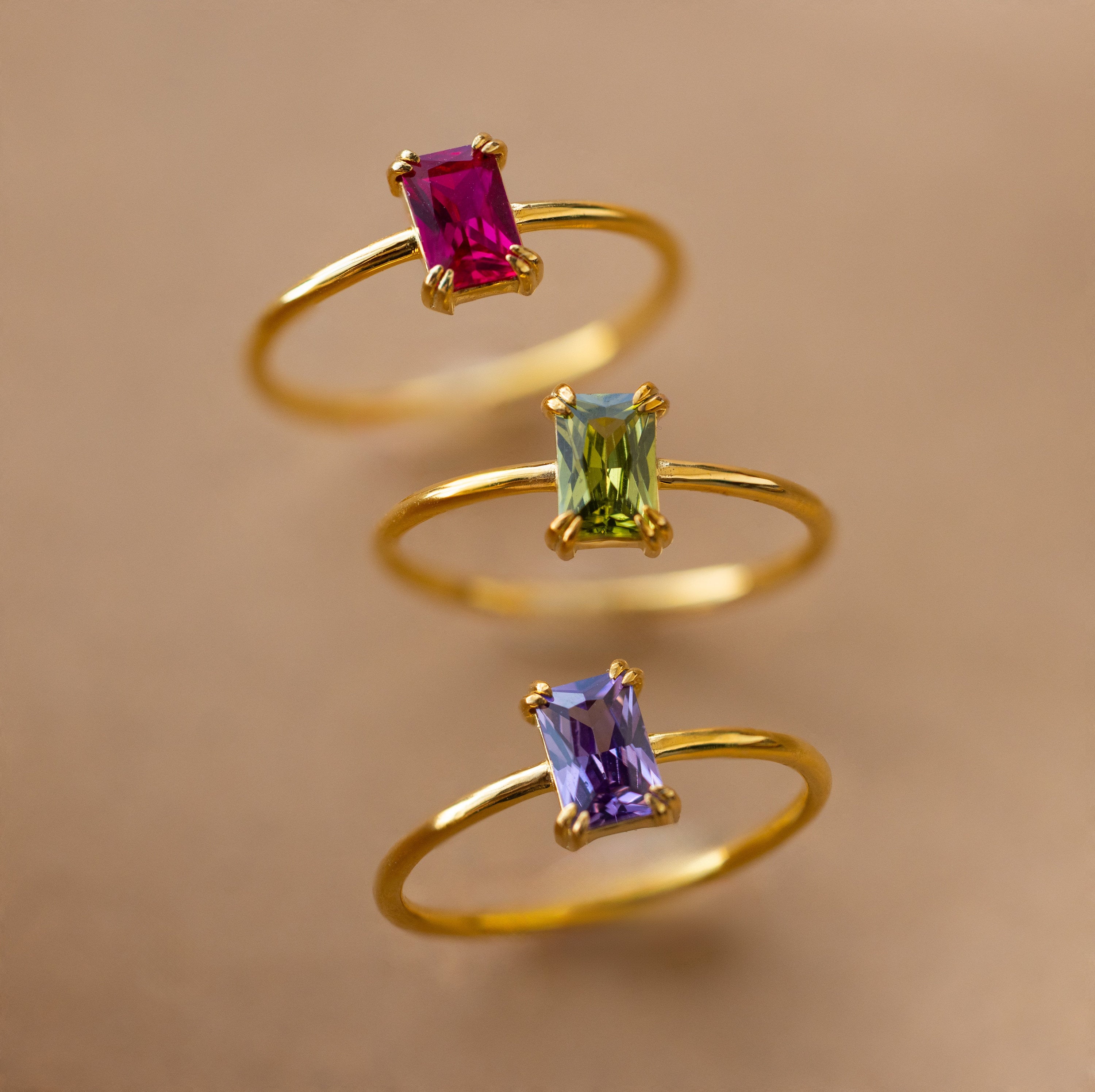 Three Emerald Cut Birthstone Rings—featuring rectangular pink, green, and purple gemstones—shine beautifully on a soft brown background. The perfect thoughtful Christmas gift.