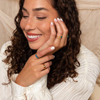 A woman with curly hair smiles, wearing a white blouse and colorful rings, including the Oval Birthstone Ring, her nails painted white.