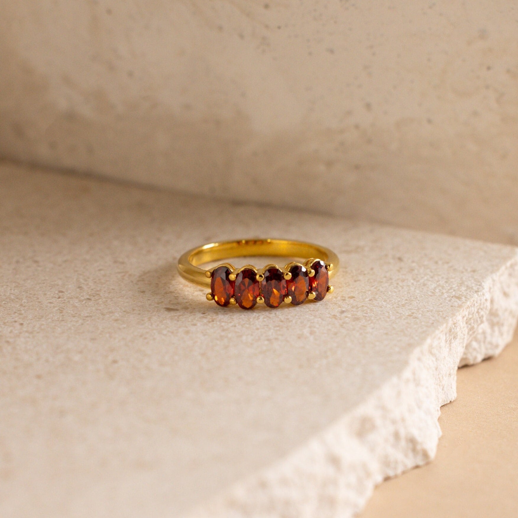 The Oval Birthstone Ring showcases five vibrant red gemstones, elegantly arranged on a textured, light-colored stone background.