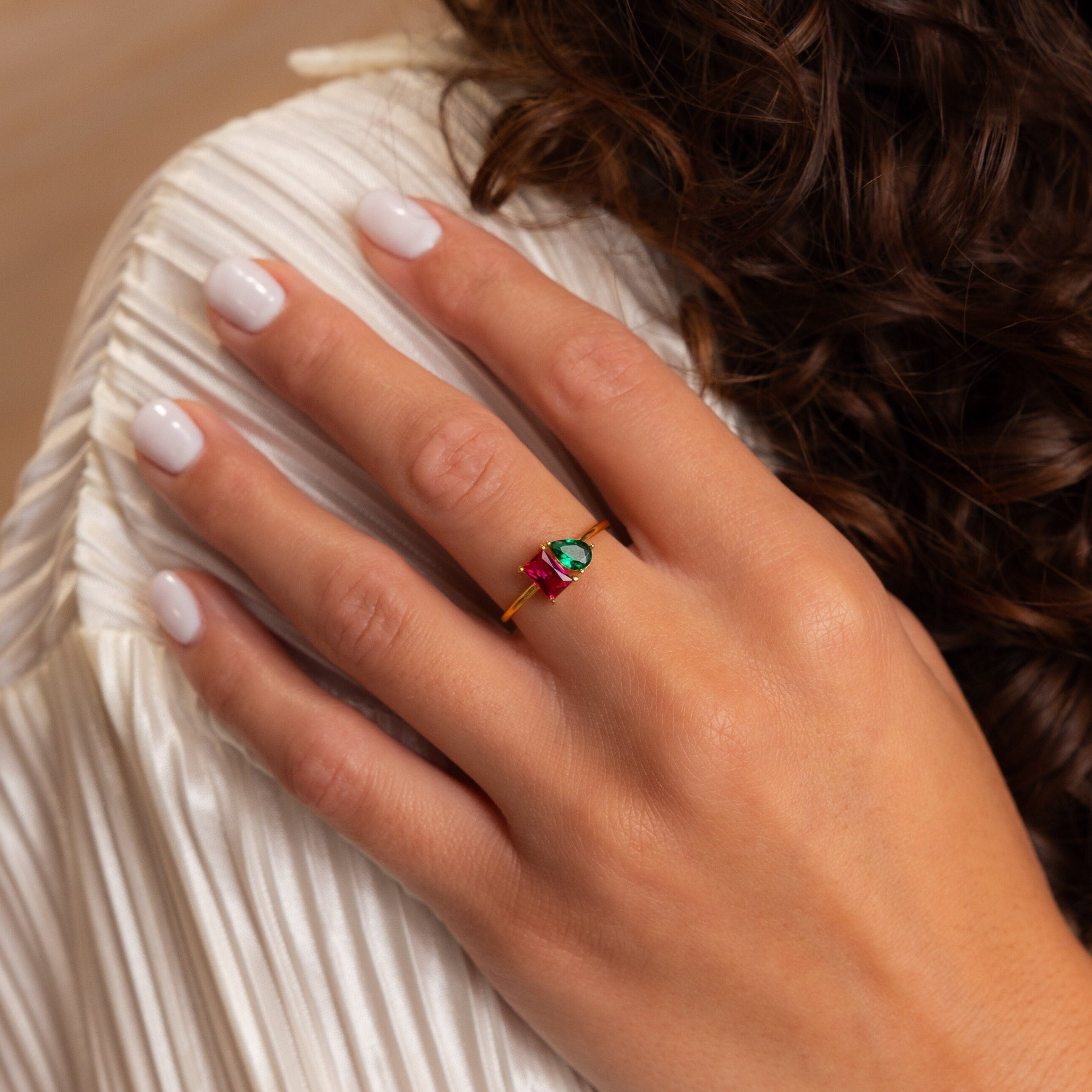 A hand with white nails wears the Toi et Moi Birthstone Ring—a gold customizable ring set with green, pink, and clear gemstones—on a white pleated sleeve, making it a perfect unique Christmas gift.