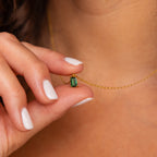 A hand with white nails holds the Emerald Cut Birthstone Necklace featuring a green pendant near the neck—an elegant, customizable piece that makes a stunning Christmas gift.
