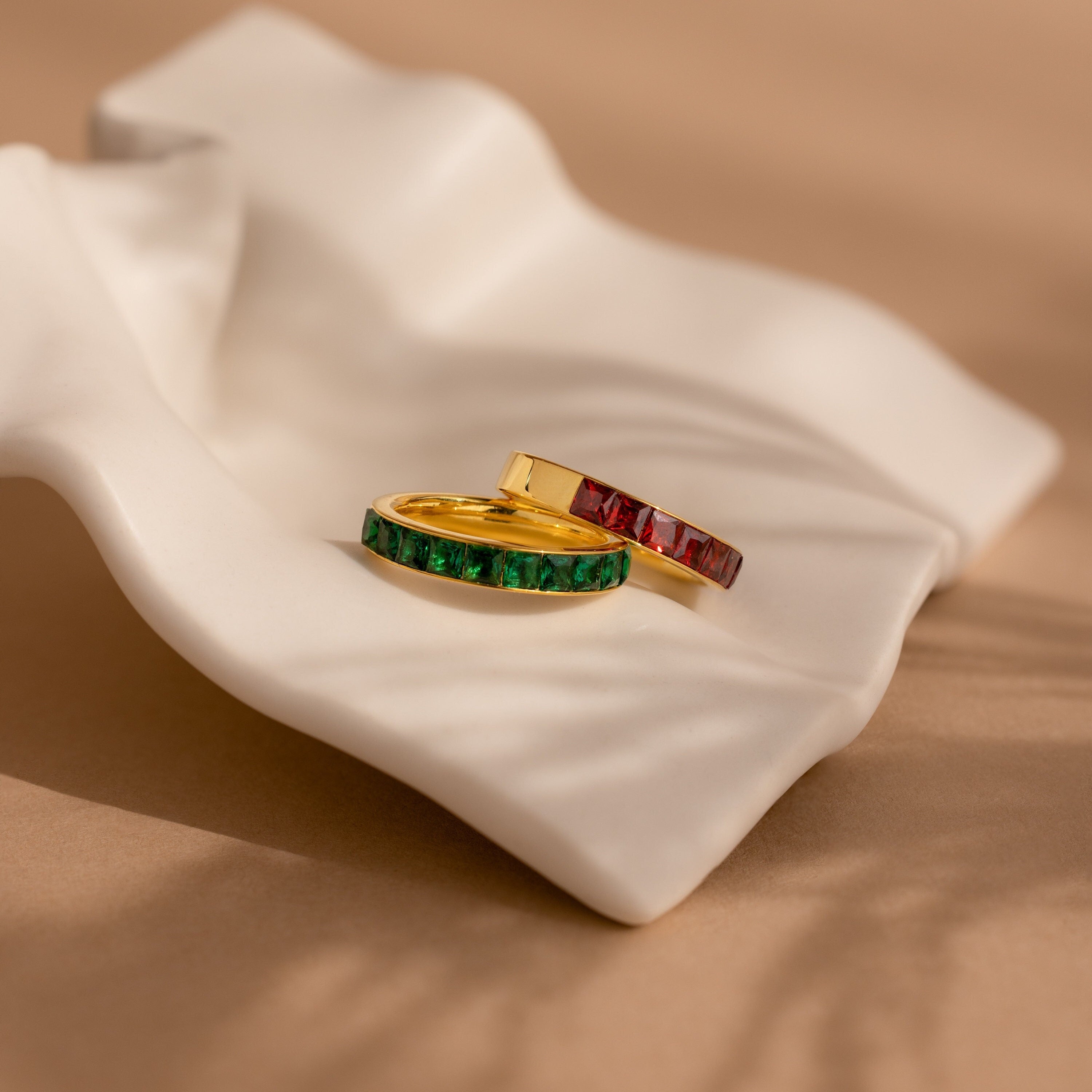 Two Birthstone Inlay Bands with green and red gemstones rest on a white, wavy ceramic dish against a beige background—a striking statement accessory for any occasion.