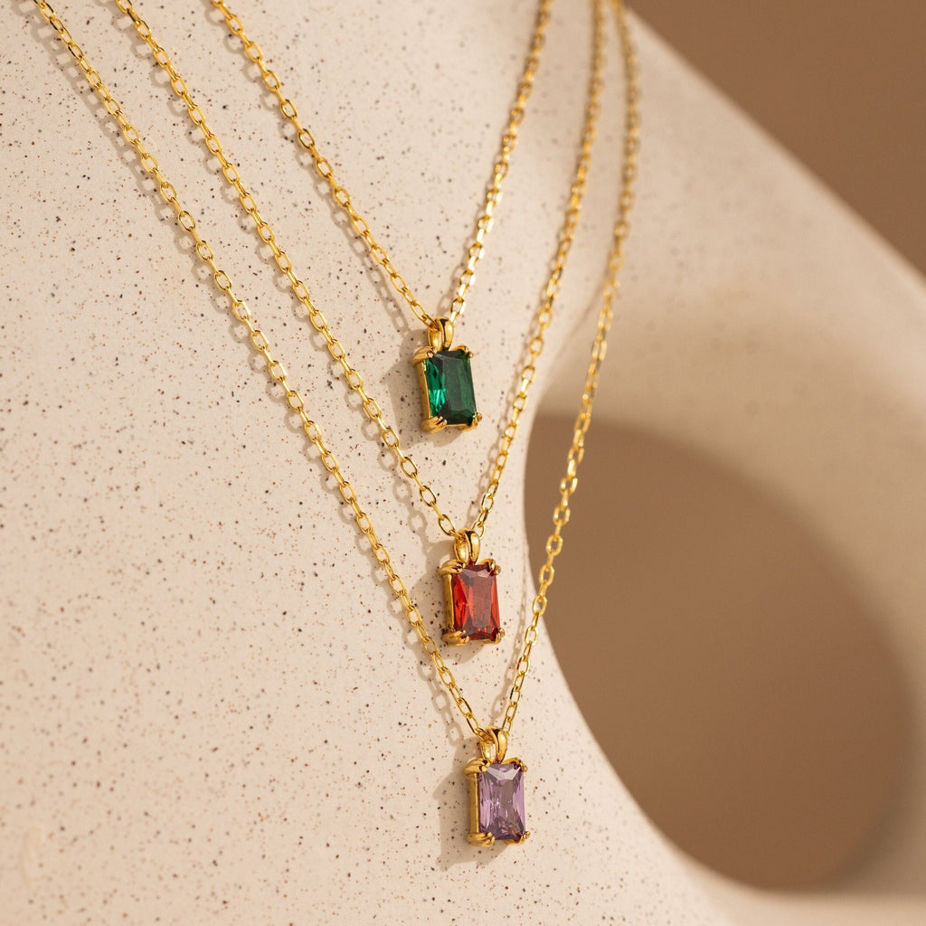 Three gold necklaces with rectangular green, red, and purple gemstone pendants rest on a beige textured surface—the perfect Emerald Cut Birthstone Necklace for a unique Christmas gift.