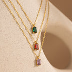 Three gold necklaces with rectangular green, red, and purple gemstone pendants rest on a beige textured surface—the perfect Emerald Cut Birthstone Necklace for a unique Christmas gift.