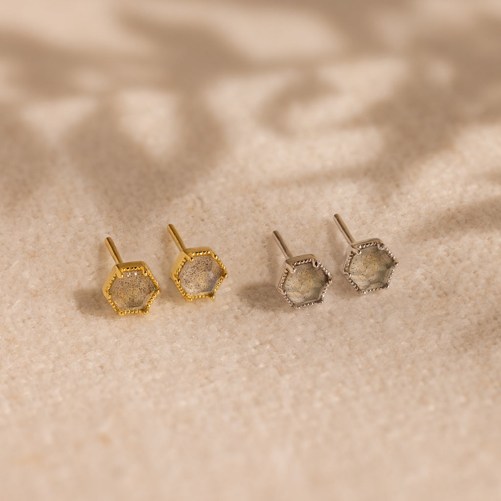A pair of Labradorite Hexagon Studs, one in gold and one in silver, displayed on a sandy beige surface—offering a subtle nod to mystical jewelry.