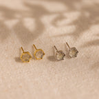 A pair of Labradorite Hexagon Studs, one in gold and one in silver, displayed on a sandy beige surface—offering a subtle nod to mystical jewelry.