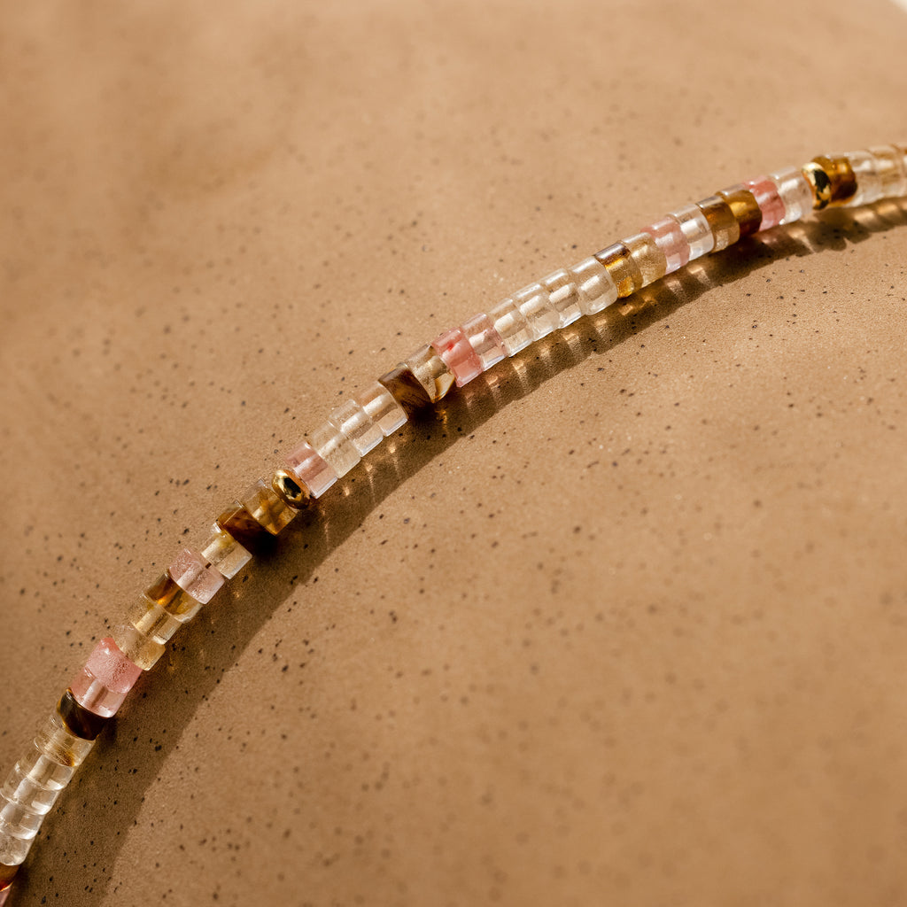 The Rose Quartz Beaded Necklace with pink, gold, and clear beads rests on a speckled beige surface—an elegant gift for your best friend.