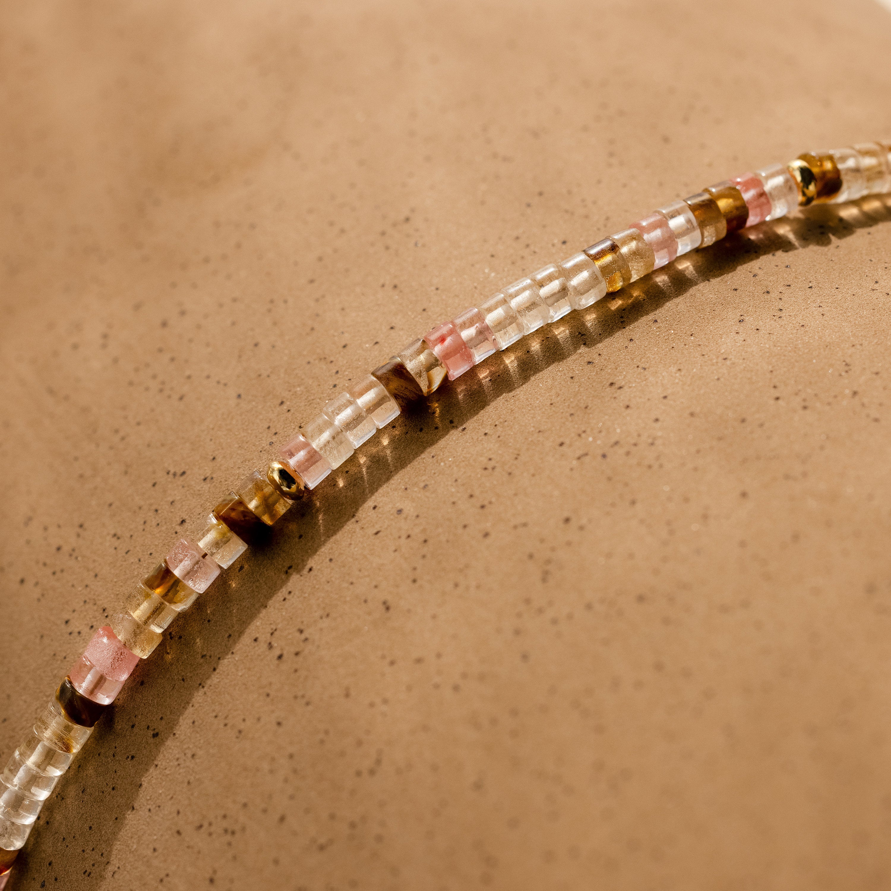 The Rose Quartz Beaded Necklace with pink, gold, and clear beads rests on a speckled beige surface—an elegant gift for your best friend.