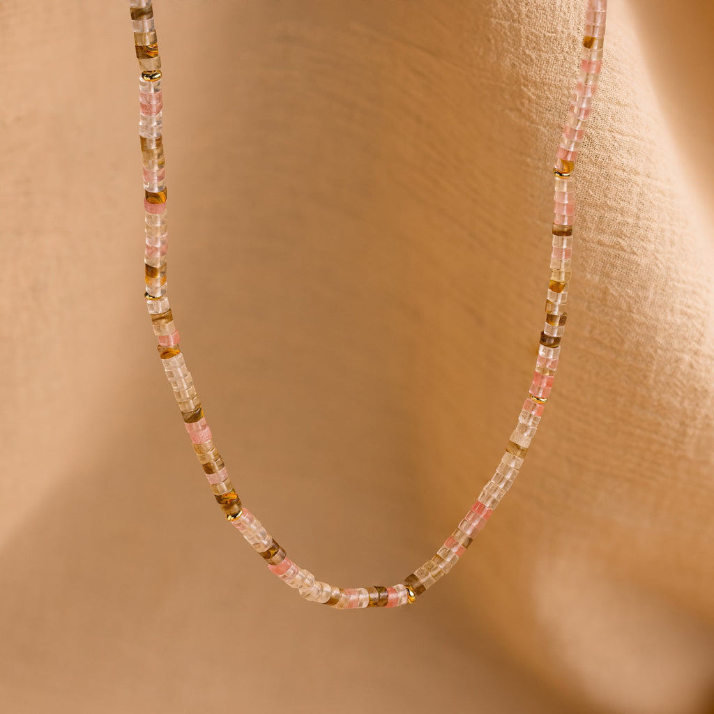The Rose Quartz Beaded Necklace features pink, clear, and gold beads and is shown on a soft beige fabric background—a thoughtful gift for your best friend.