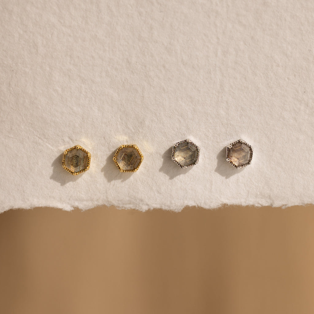 Add a mystical touch with the Labradorite Hexagon Studs—this set includes four earrings, two gold and two silver, displayed on a textured off-white surface. Perfect for those who adore unique hexagon-shaped jewelry.