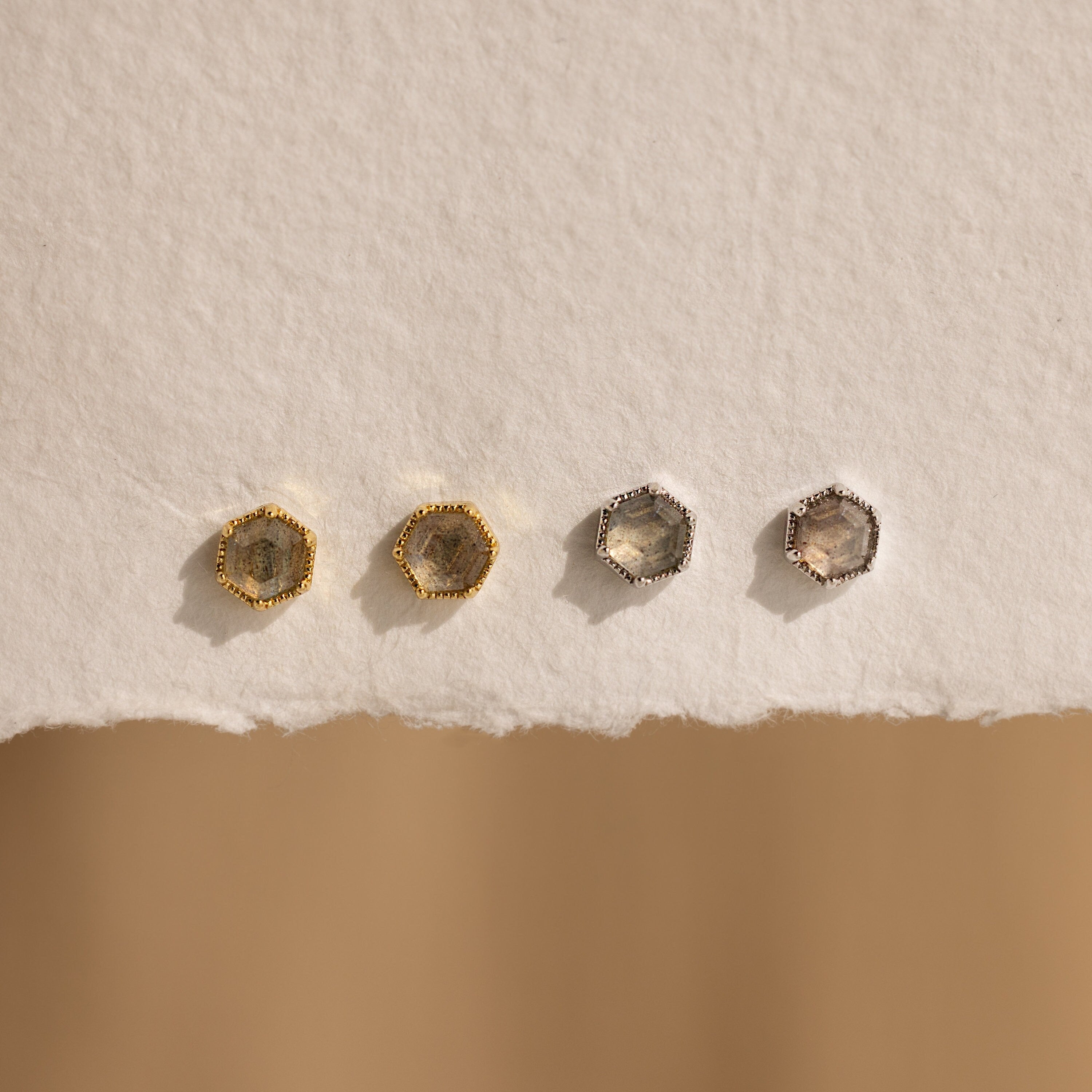 Add a mystical touch with the Labradorite Hexagon Studs—this set includes four earrings, two gold and two silver, displayed on a textured off-white surface. Perfect for those who adore unique hexagon-shaped jewelry.