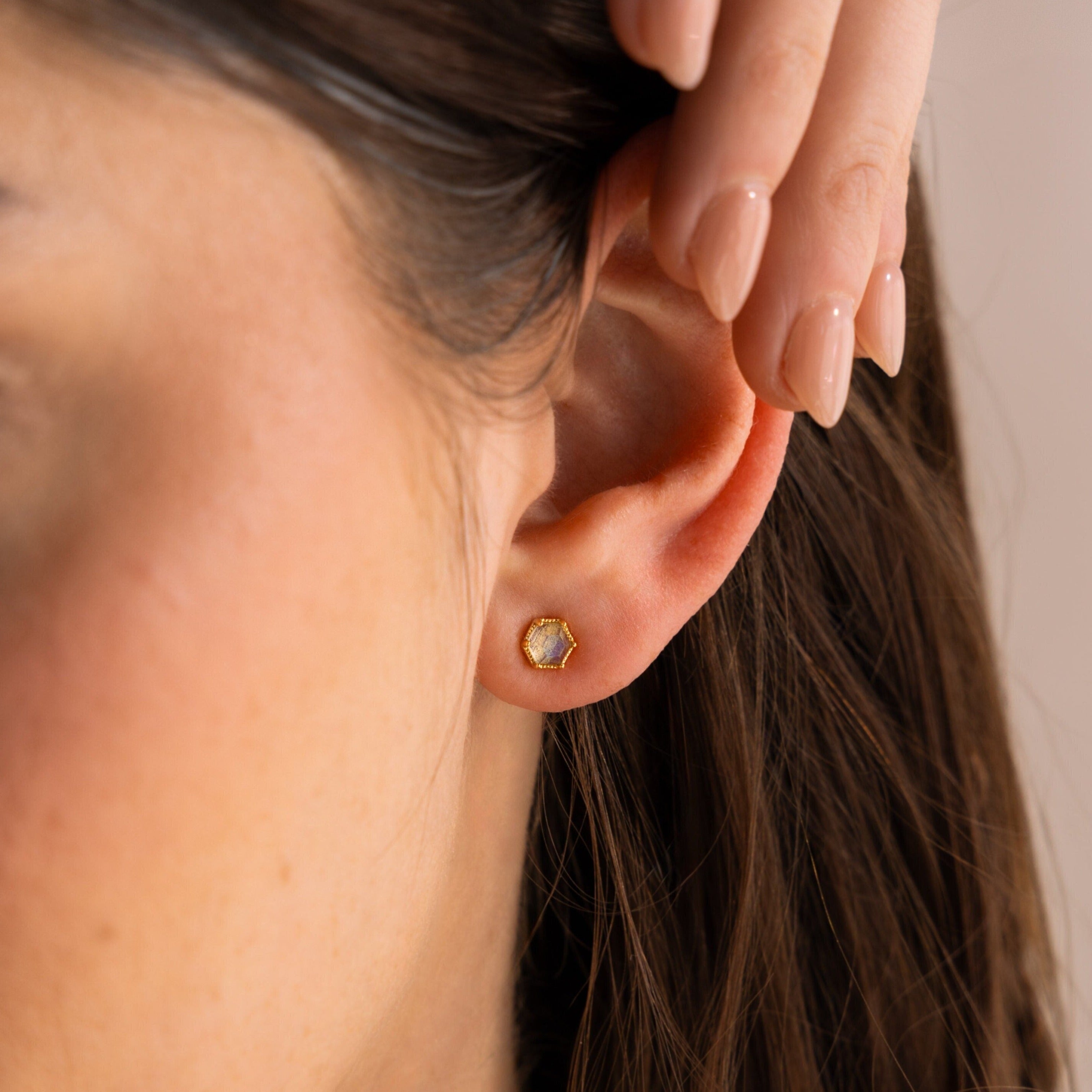 A woman’s ear adorned with Labradorite Hexagon Studs—a gold, hexagonal earring—her hand gently touching her head.