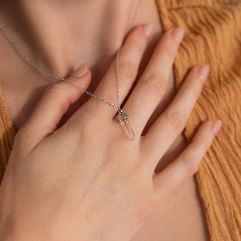 A hand touches a Clear Quartz Necklace with a hexagonal point pendant, worn over a textured, light brown top.