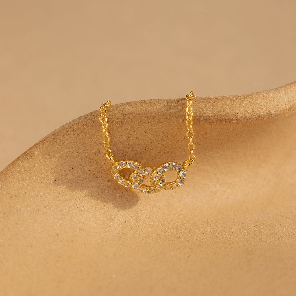 The Pave Link Necklace showcases two diamond-studded interlocking links on a gold chain, elegantly presented on a beige surface—a stunning addition to any jewelry collection.
