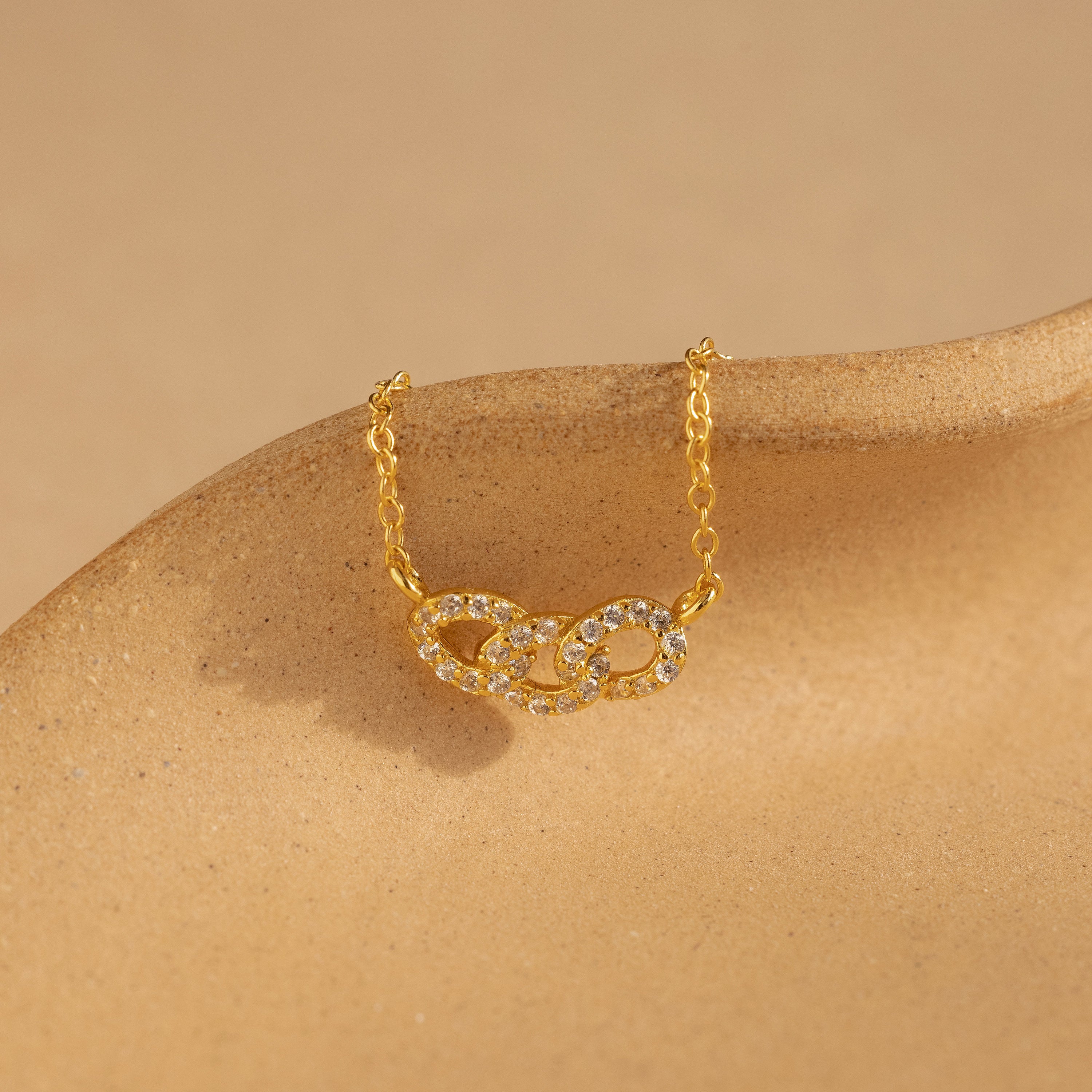The Pave Link Necklace showcases two diamond-studded interlocking links on a gold chain, elegantly presented on a beige surface—a stunning addition to any jewelry collection.