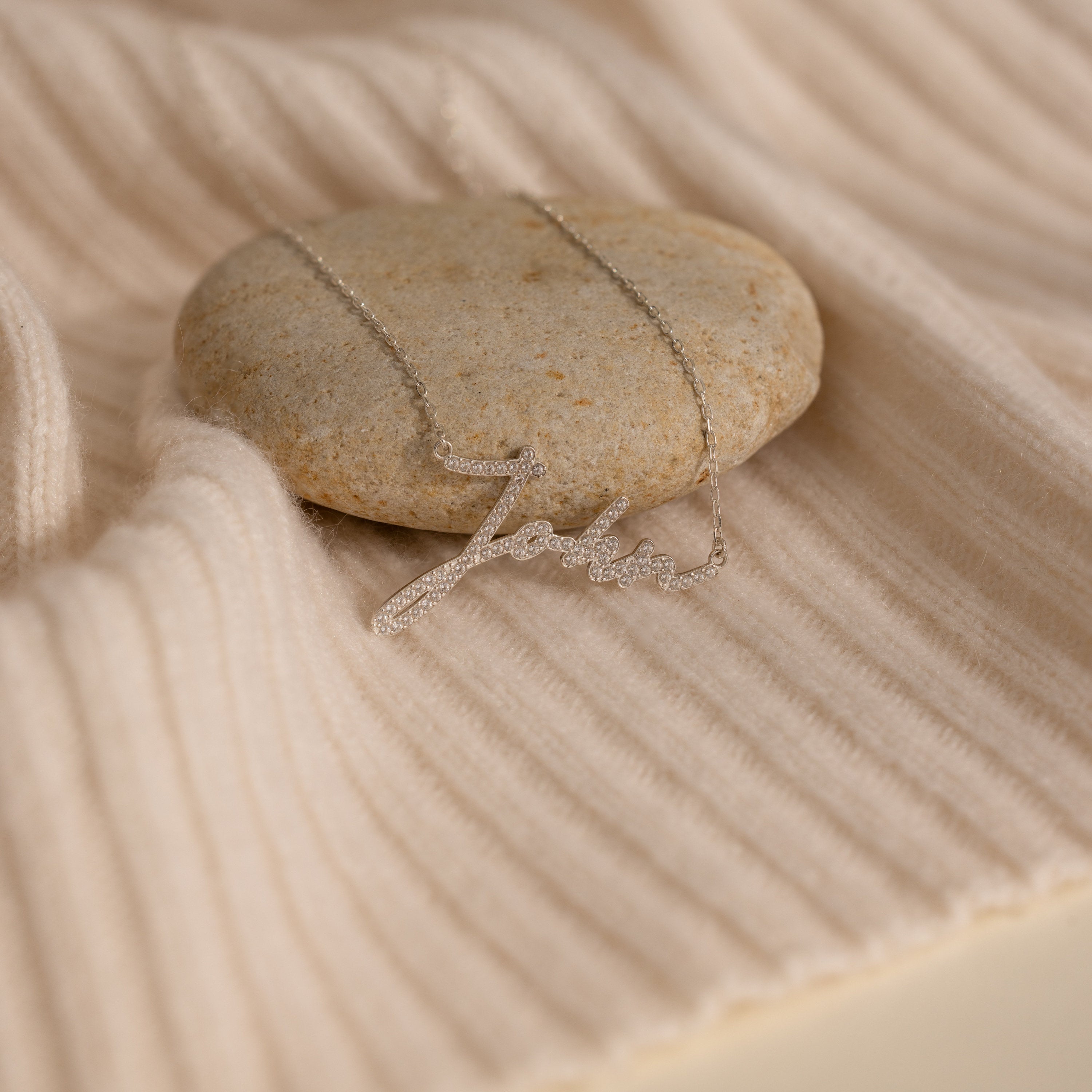 The Pave Handwriting Necklace rests on a smooth stone and cream ribbed fabric, highlighting its timeless statement jewelry style.