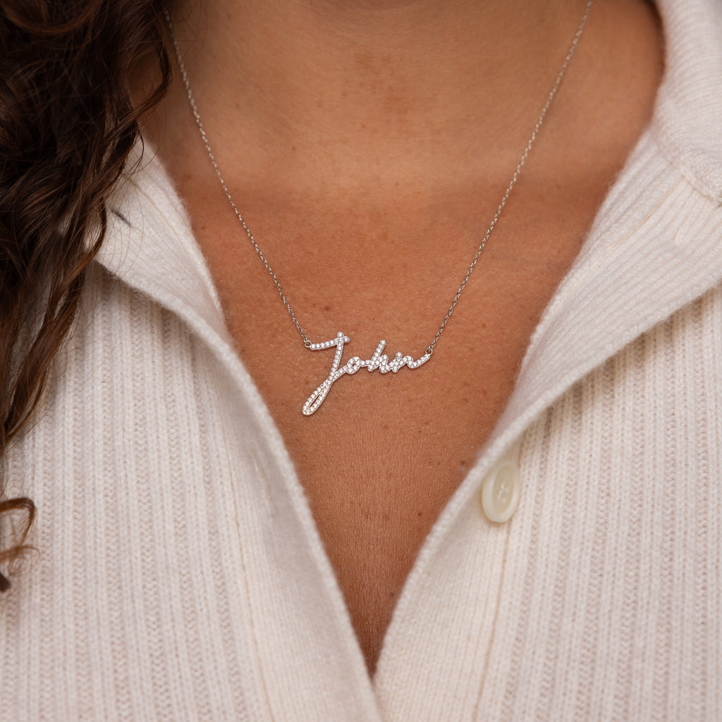 A woman wears a white sweater and the Pave Handwriting Necklace featuring the name Julia in elegant cursive—a striking statement piece.