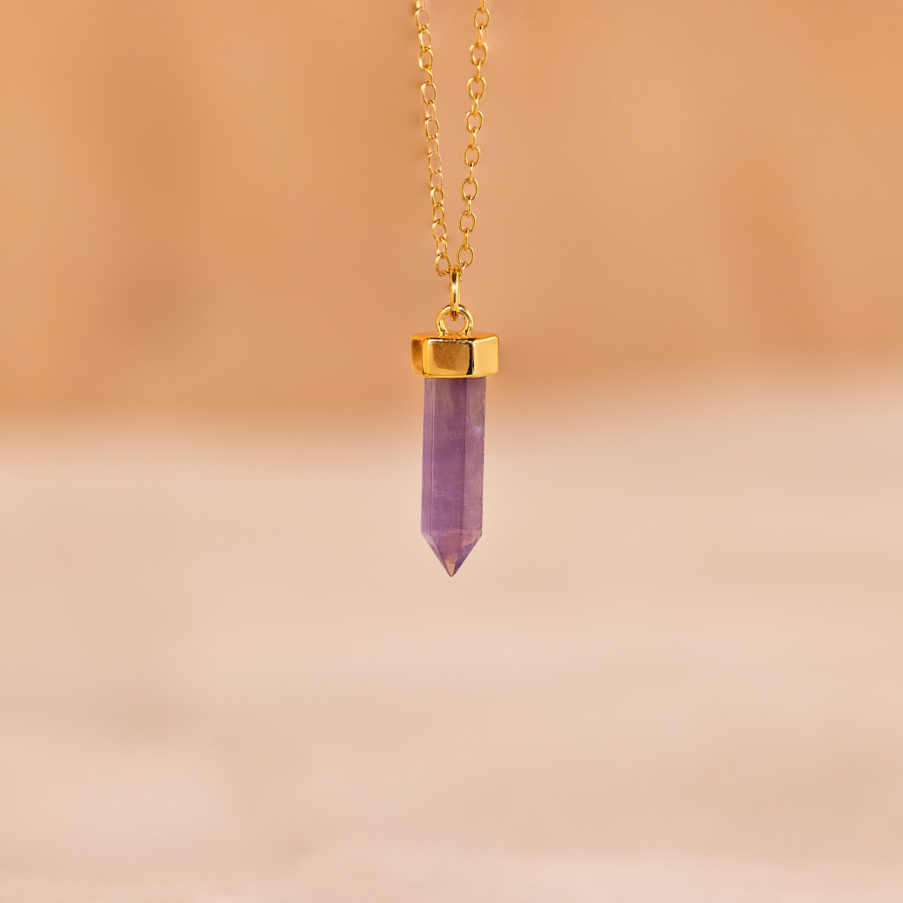 The Amethyst Quartz Necklace features a gold chain with a crystal charm pendant, showcased against a soft beige background—a unique gift for any occasion.