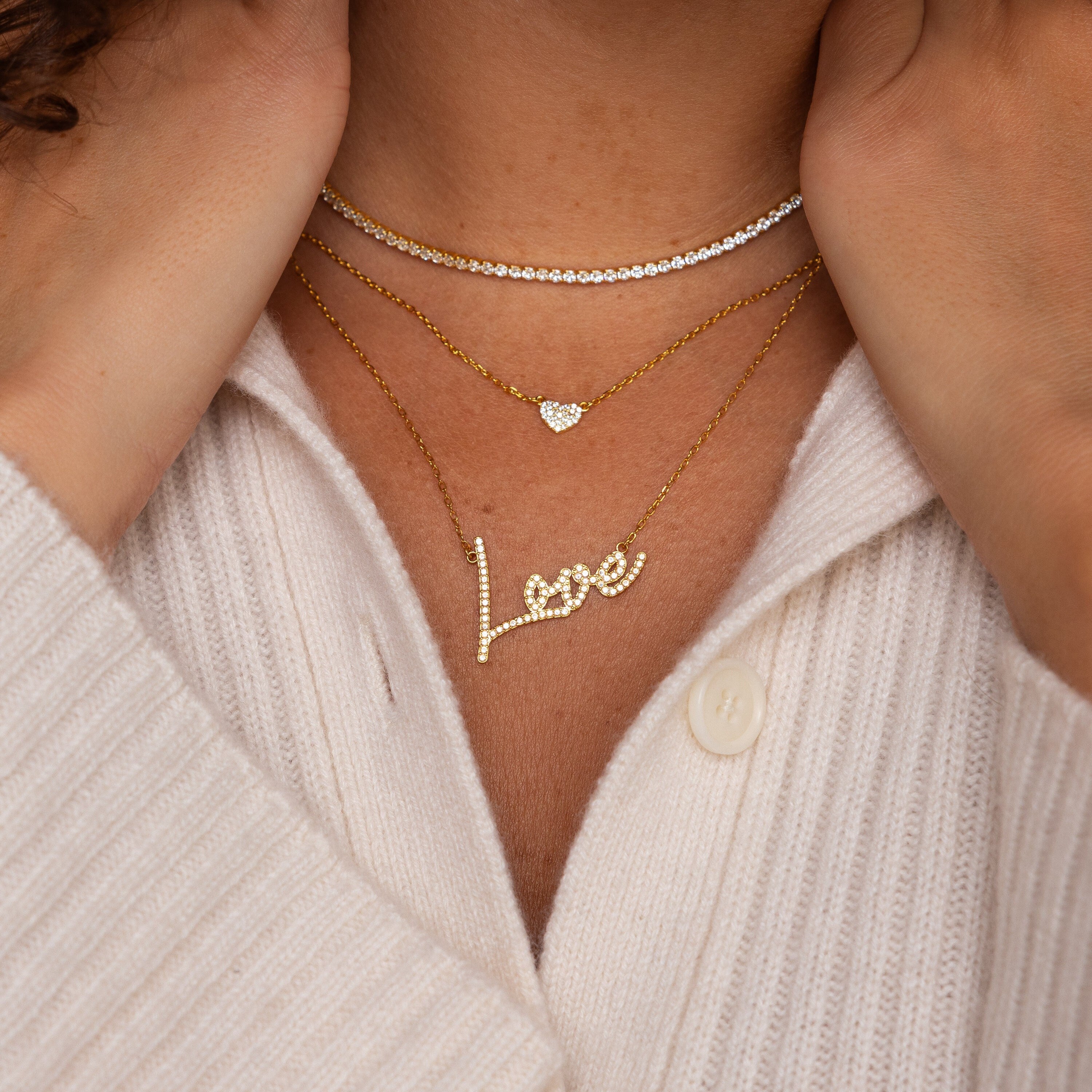 A person wears the Pave Handwriting Necklace, spelling "Love," layered with gold necklaces over a cream ribbed sweater for an effortlessly chic statement jewelry look.