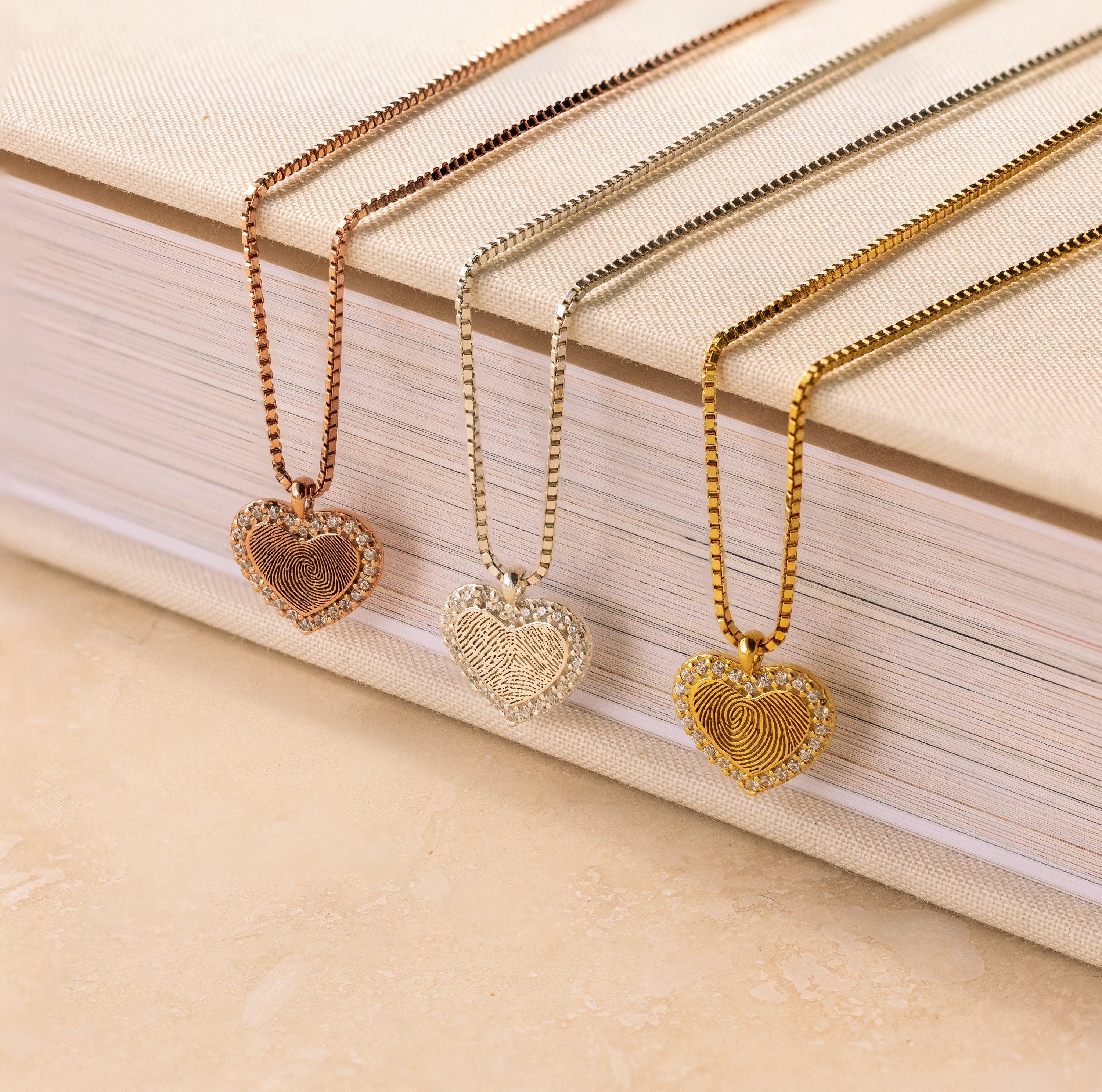 Three Pave Fingerprint Heart Necklaces in rose gold, silver, and gold sit on a beige-covered book—each personalized with a fingerprint for a unique touch.