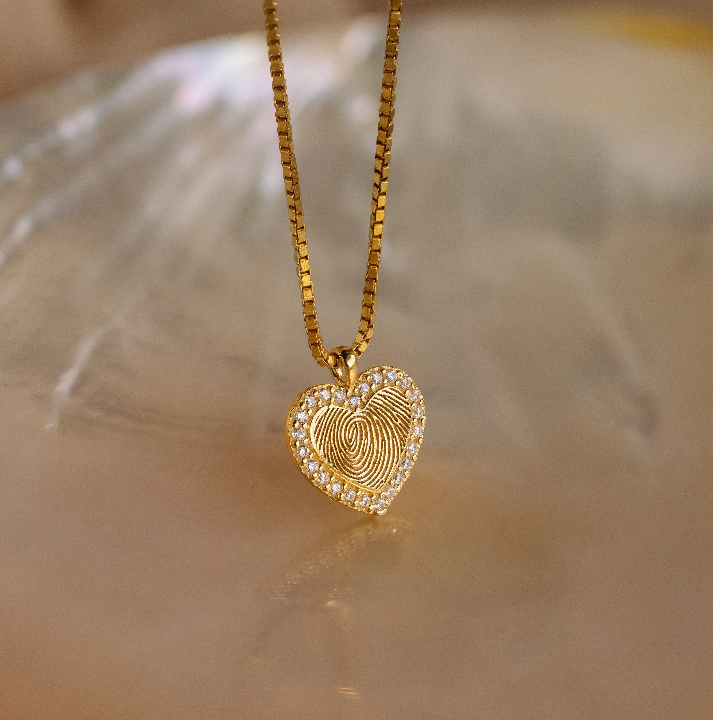 The Pave Fingerprint Heart Necklace is a gold heart-shaped pendant featuring your engraved fingerprint, accented with small gems, and hung on a gold chain—a beautiful personalized jewelry piece.