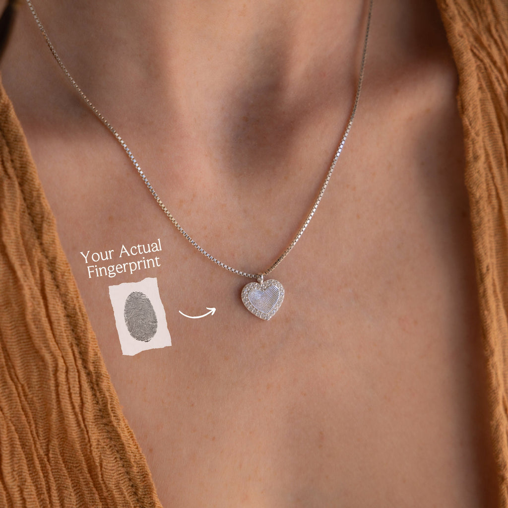 The Pave Fingerprint Heart Necklace, featuring an engraved fingerprint heart pendant, rests on a person's neck and is displayed with a sample fingerprint image and label.