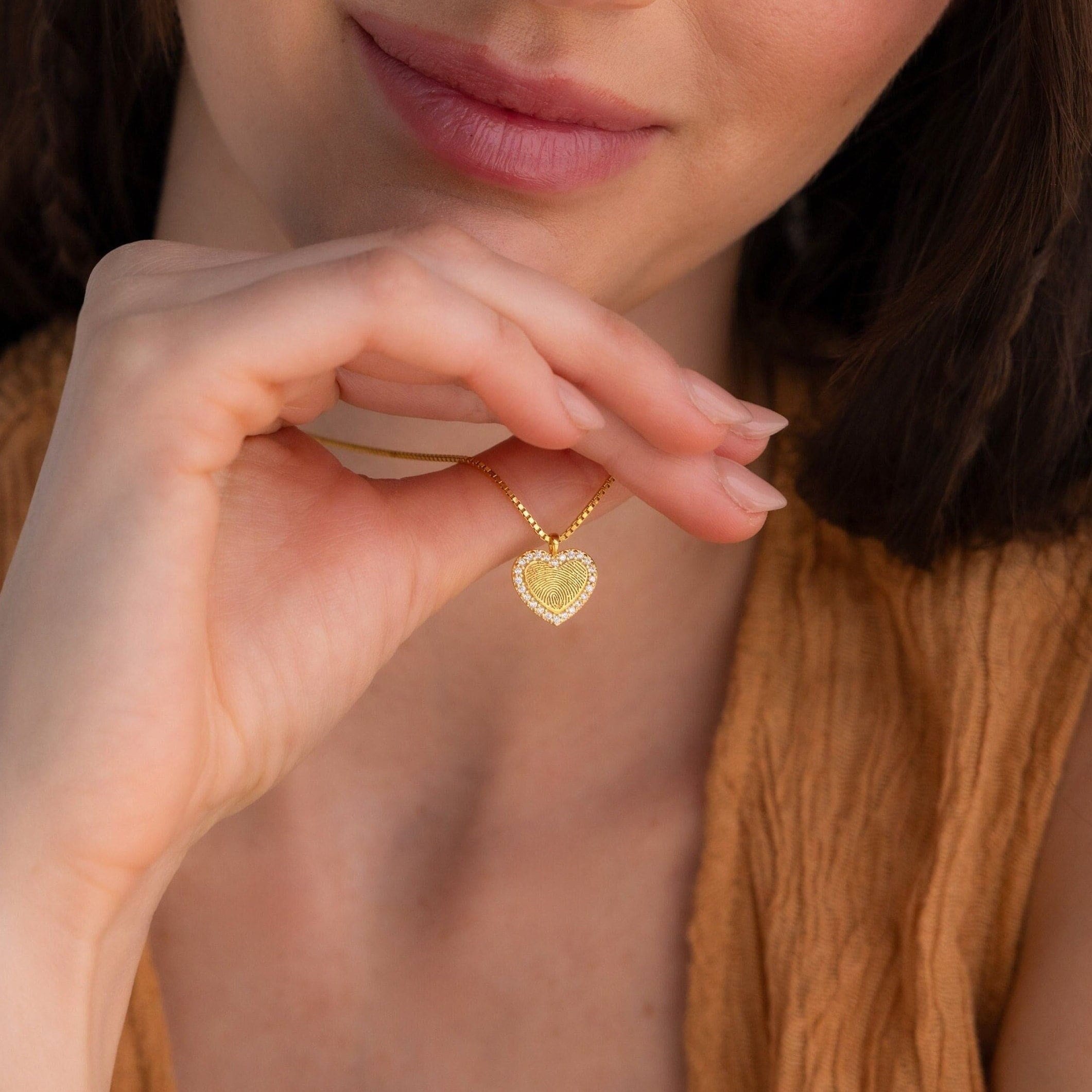 A woman wearing a brown top holds a personalized Pave Fingerprint Heart Necklace close to her face.