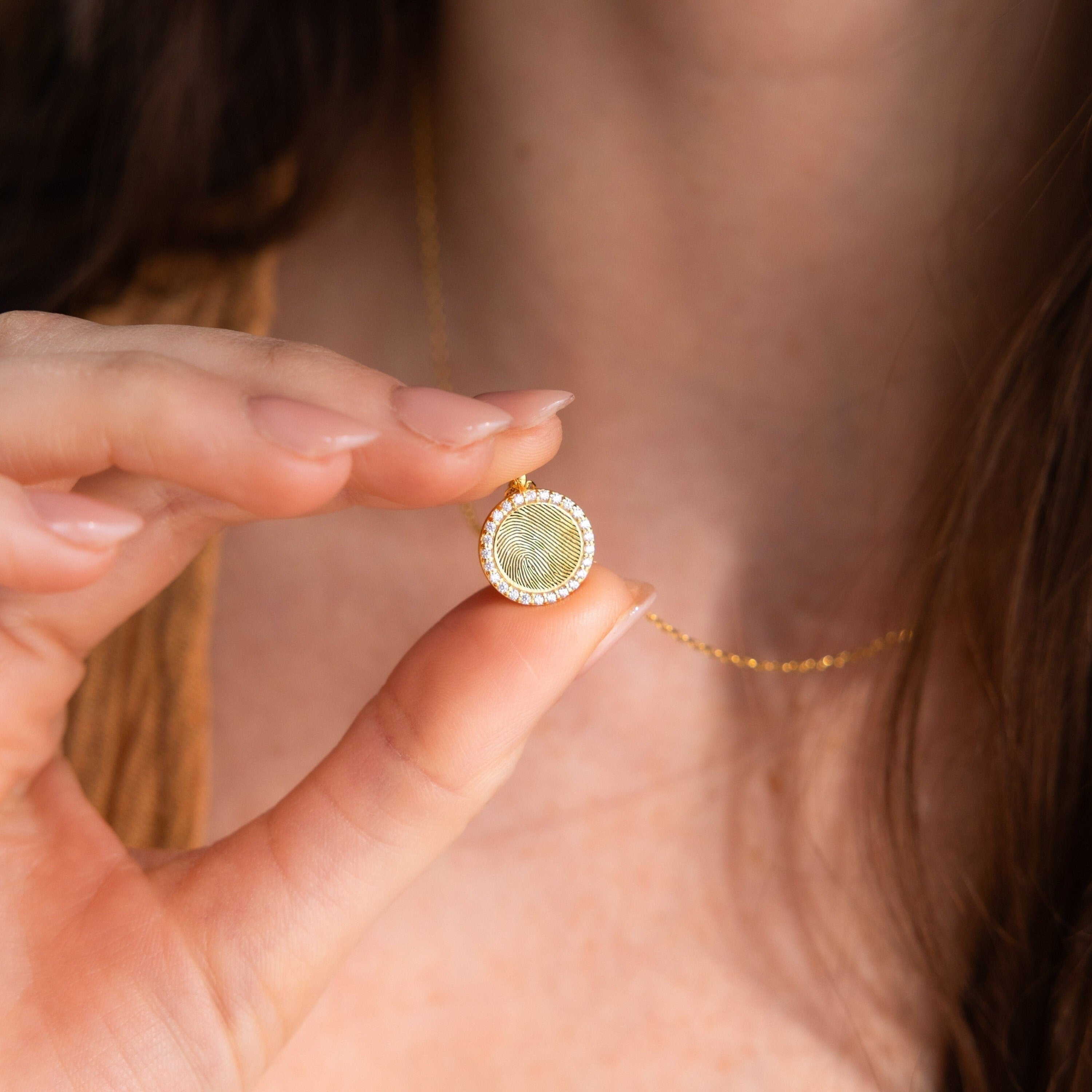 A person holding a gold Pave Fingerprint Coin Necklace close to their neck—an elegant engraved pendant perfect as a heartfelt memorial gift.