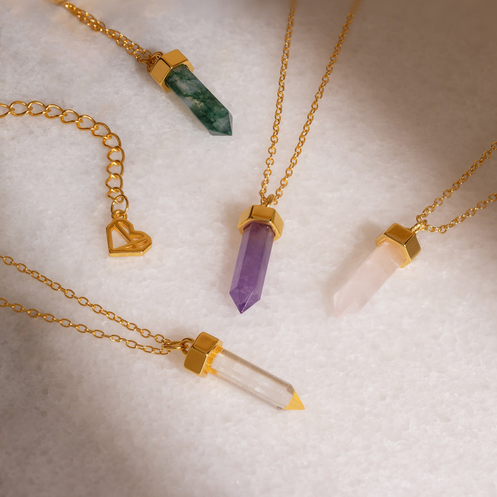The Agate Quartz Necklace, featuring a gold chain and green crystal pendant, shines alongside other green, purple, pink, and clear crystal necklaces on a white surface.