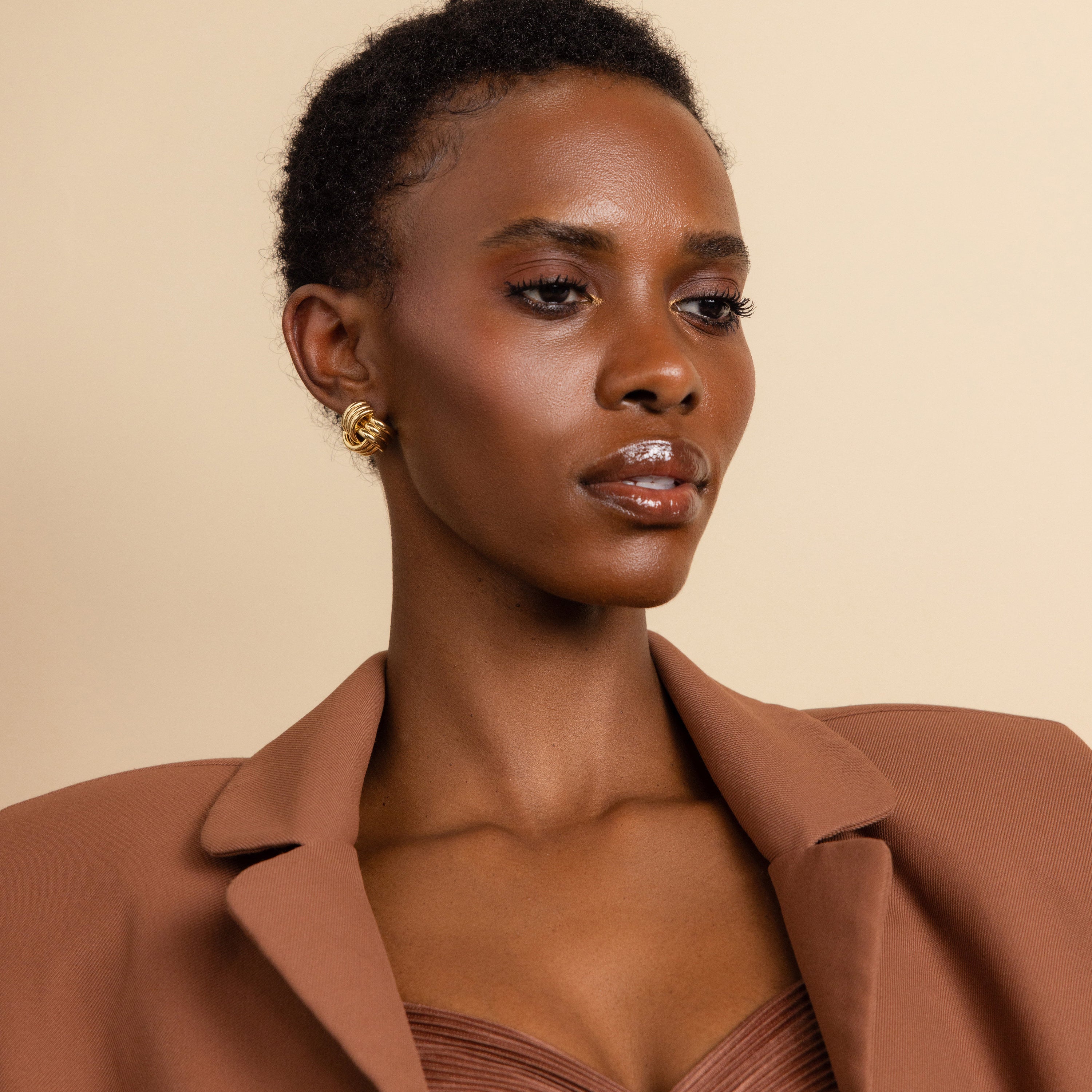 A woman with short hair, dressed in a brown blazer, showcases the Bold Triple Knot Studs—rope-inspired earrings—while posing against a beige background.