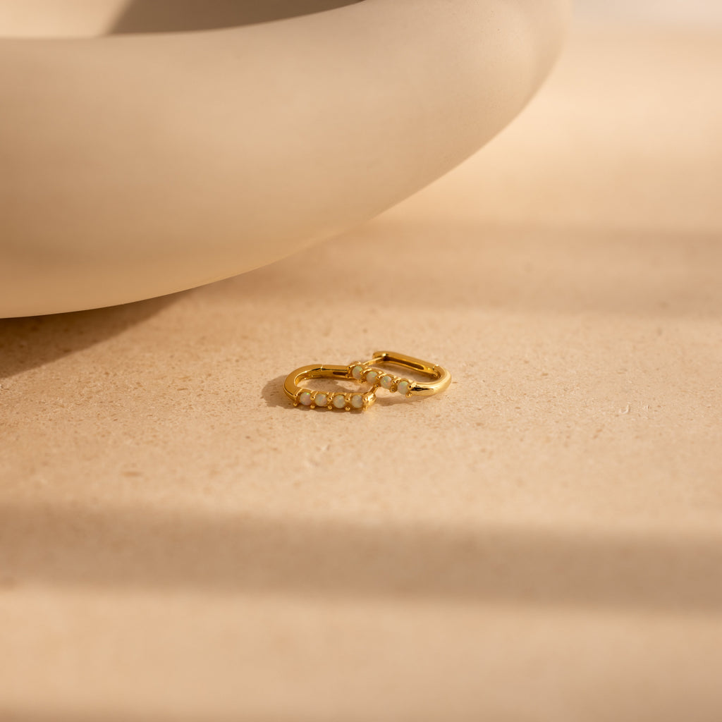 A pair of Opal Link Hoops with small pearls are displayed on a beige surface beside a curved ceramic piece.