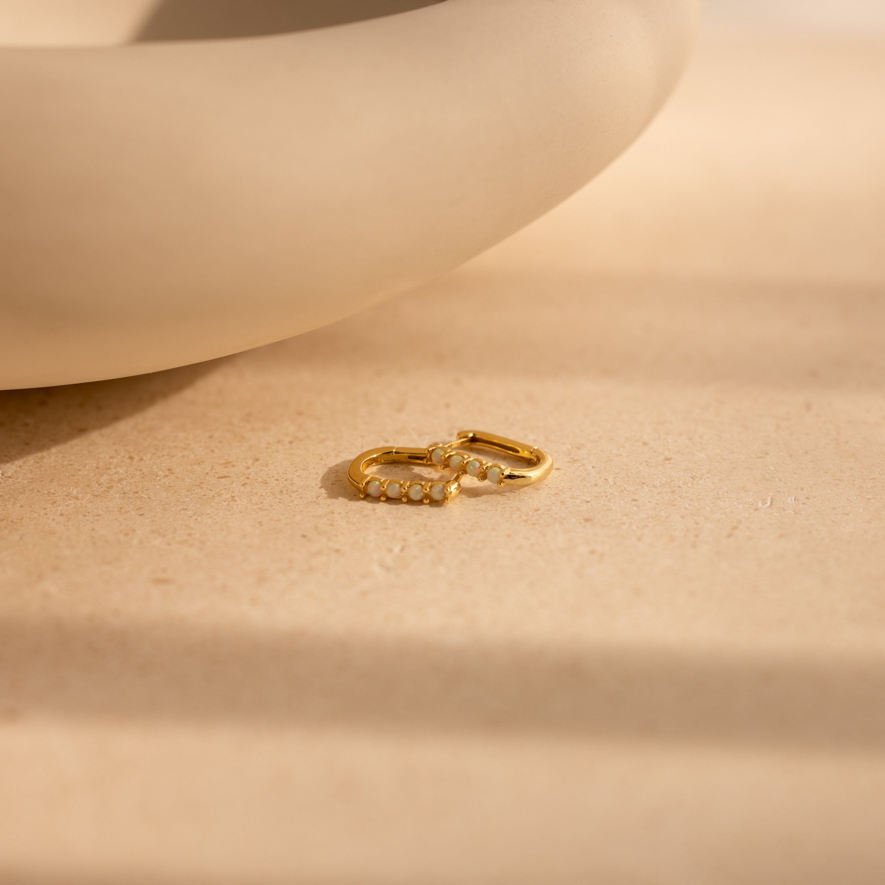 A pair of Opal Link Hoops with small pearls are displayed on a beige surface beside a curved ceramic piece.