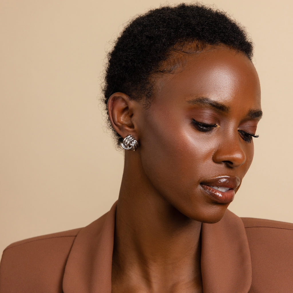 A woman looks down in a brown jacket, wearing Bold Triple Knot Studs that add subtle detail to her look.