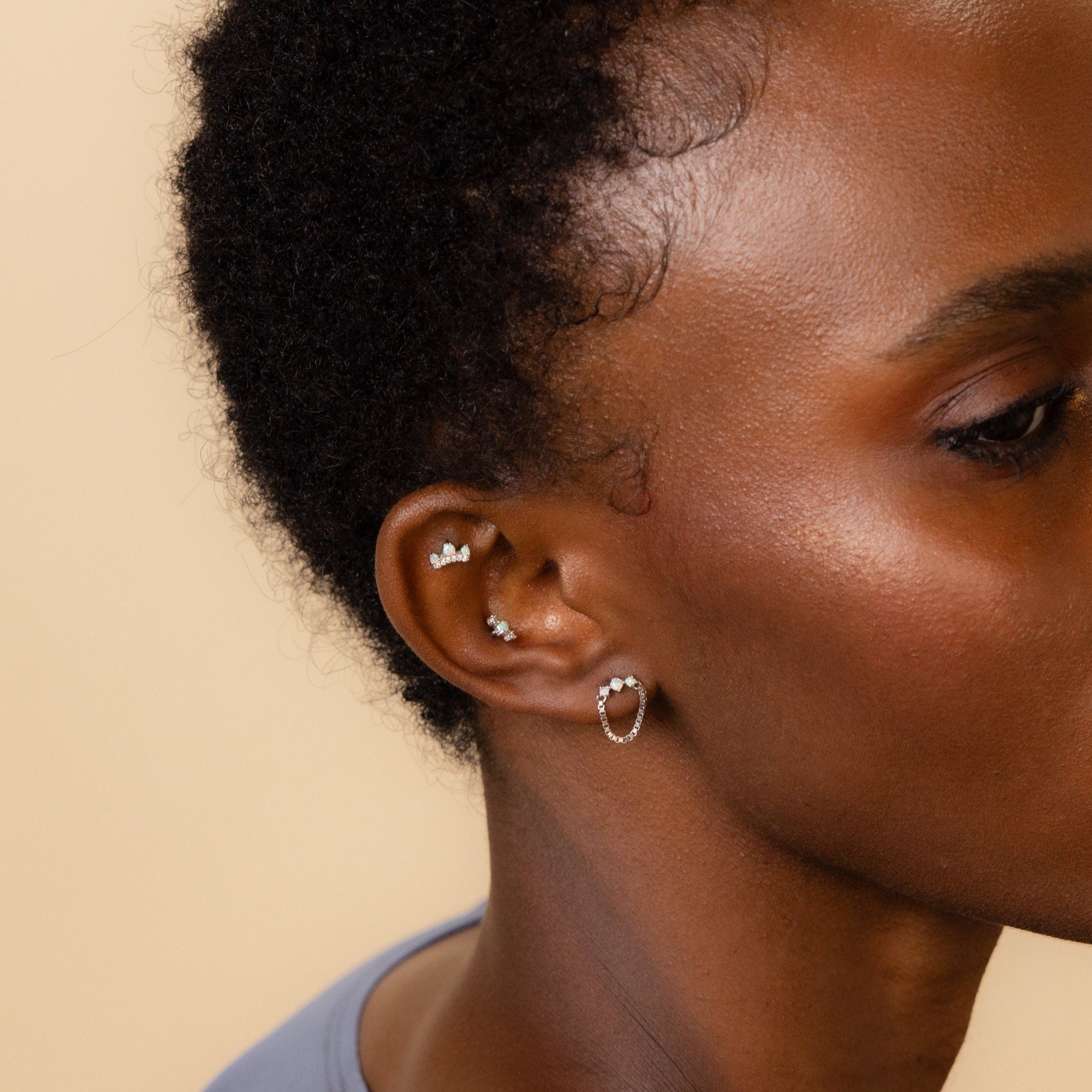 Close-up of a person’s ear adorned with multiple silver earrings, including hoops, Helix piercings, and the Opal Cartilage Studs Set, against a beige background.